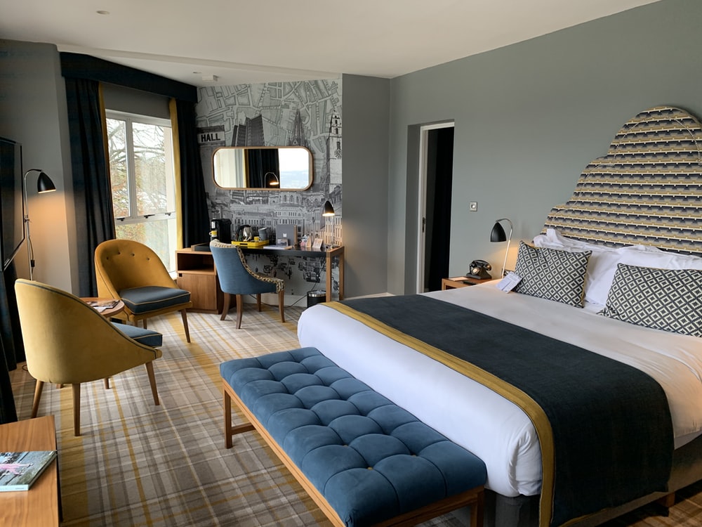 Elegant boutique hotel room interior with a large bed, blue upholstered bench, mustard armchairs, and decorative wallpaper featuring architectural elements.